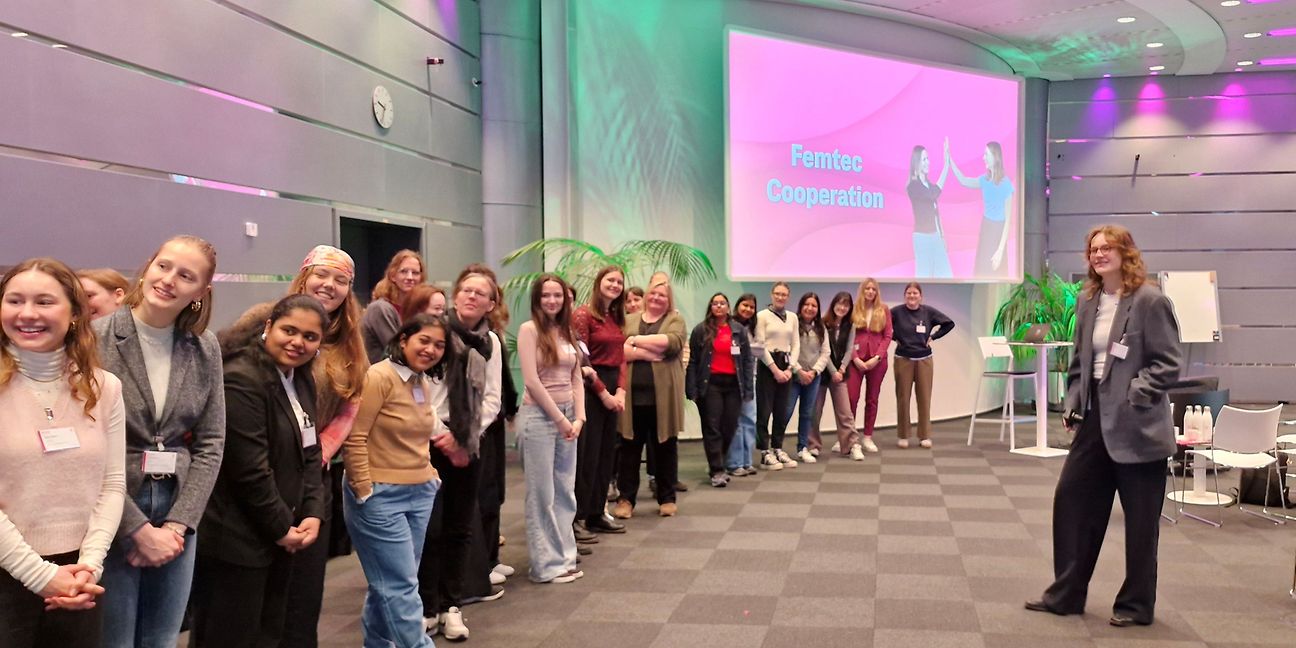Junge MINT Studentinnen stehen vor einem Präsentations-Bildschirm auf dem „Femtec Cooperation“ steht