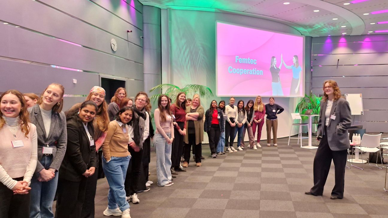 Young female STEM students stand in front of a presentation screen that reads “Femtec Cooperation”