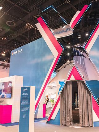 Eingangsbereich auf der DIGITAL X Messe mit großem Pavillon in X-Form, an dem Menschen vorbeigehen und hineingehen.