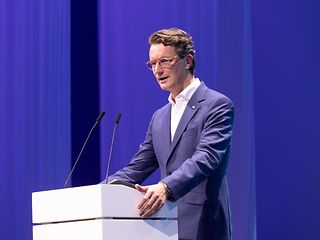 Hendrik Wüst im blauen Anzug steht auf einer Bühne hinter einem weißen Rednerpult und spricht in zwei Mikrofone.