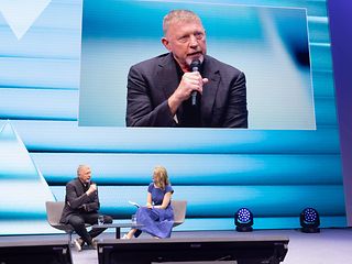 Boris Becker is sitting on stage with a microphone, giving a talk.