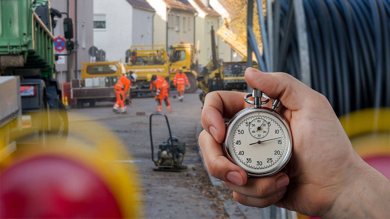 Hand hält analoge Stoppuhr. Im Hintergrund eine Baustelle mit Arbeitern