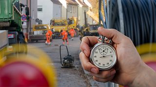 Hand hält analoge Stoppuhr. Im Hintergrund eine Baustelle mit Arbeitern