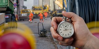 Hand hält analoge Stoppuhr. Im Hintergrund eine Baustelle mit Arbeitern