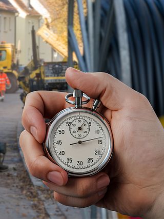 Hand hält analoge Stoppuhr. Im Hintergrund eine Baustelle mit Arbeitern