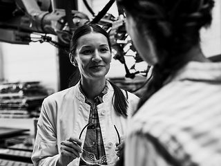 Eine freundliche junge Frau mit einer Schutzbrille in den Händen steht vor einer Maschine in einer Produktionshalle.