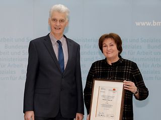 Staatssekretär Rolf Schmachtenberg überreicht Birgit Klesper die Auszeichnung. Es ist eine gerahmte Urkunde.