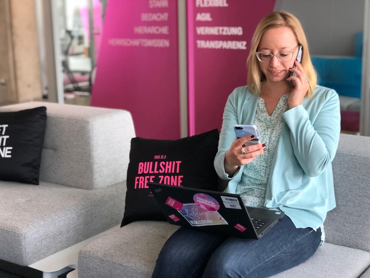 Frau sitzt auf Sofa mit Handy in der Hand, Handy am Ohr und Laptop auf dem Schoß.