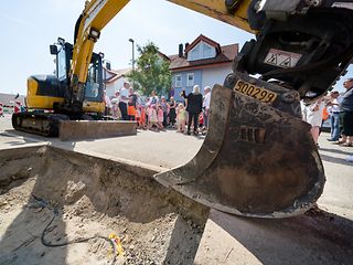 Ein gelber Bagger steht an einer Baustelle. Im Hintergrund sieht man Bürger und Kinder der Gemeinde Heimerdingen.