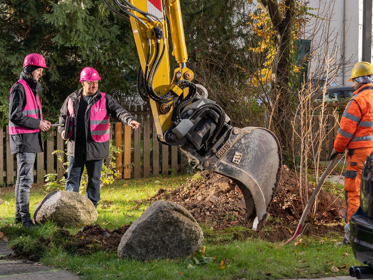 Zwei Telekom-Mitarbeiter in magenta Bauwesten stehen unter einer Baggerschaufel und sprechen über ein Ausbauvorhaben.