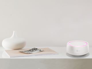 The Smart Speaker Mini in white stands on a bright living room furniture. Its illuminated ring shines in the colour magenta.