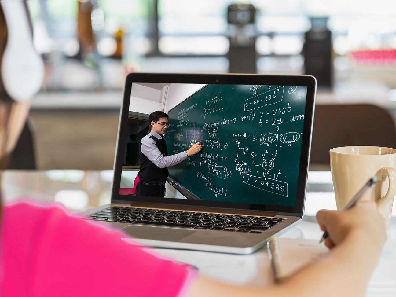 Kind sitzt mit Laptop am Tisch. Im Laptop ist zu sehen, wie ein Lehrer an der Tafel per Livestream unterrichtet.