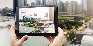 Hände, die ein Tablet halten. Das Tablet zeigt relevante Orte in der Stadt auf, während im Hintergrund die Stadt liegt.