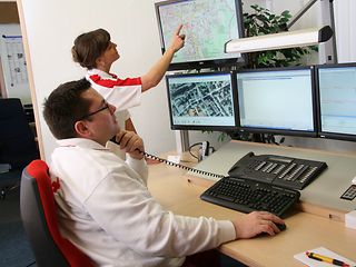 Zwei Mitarbeitende des DRK in Herten beobachten eine Wand mit Monitoren. 