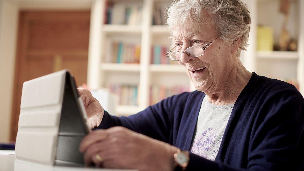 Eine ältere Dame sitzt vor einem Tablet und freut sich.