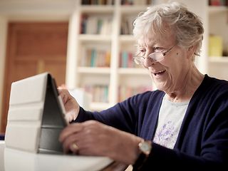 Eine ältere Dame sitzt vor einem Tablet und freut sich.