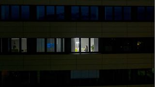 Ein dunkles Bürogebäude mit einem erleuchteten Fenster, in dem eine Frau am Rechner sitzt.