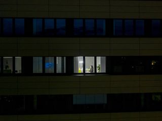 Ein dunkles Bürogebäude mit einem erleuchteten Fenster, in dem eine Frau am Rechner sitzt.