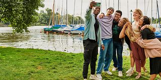 Eine Gruppe junger Menschen macht ein Selfie vor einer ländlichen Kulisse am See. 