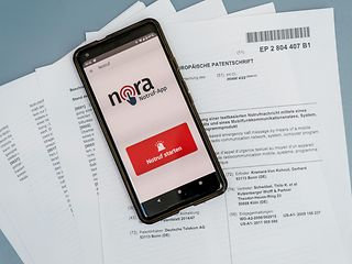  Die “Europäische Patentschrift“ auf Papier, darauf ein Smartphone mit der geöffneten Notruf-App.