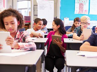 Mehrere Kinder sitzen im Klassenzimmer, die Lehrerin steht in der Mitte. Sie arbeiten mit Tablets in der Hand.