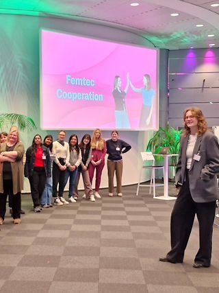 Young female STEM students stand in front of a presentation screen that reads “Femtec Cooperation”